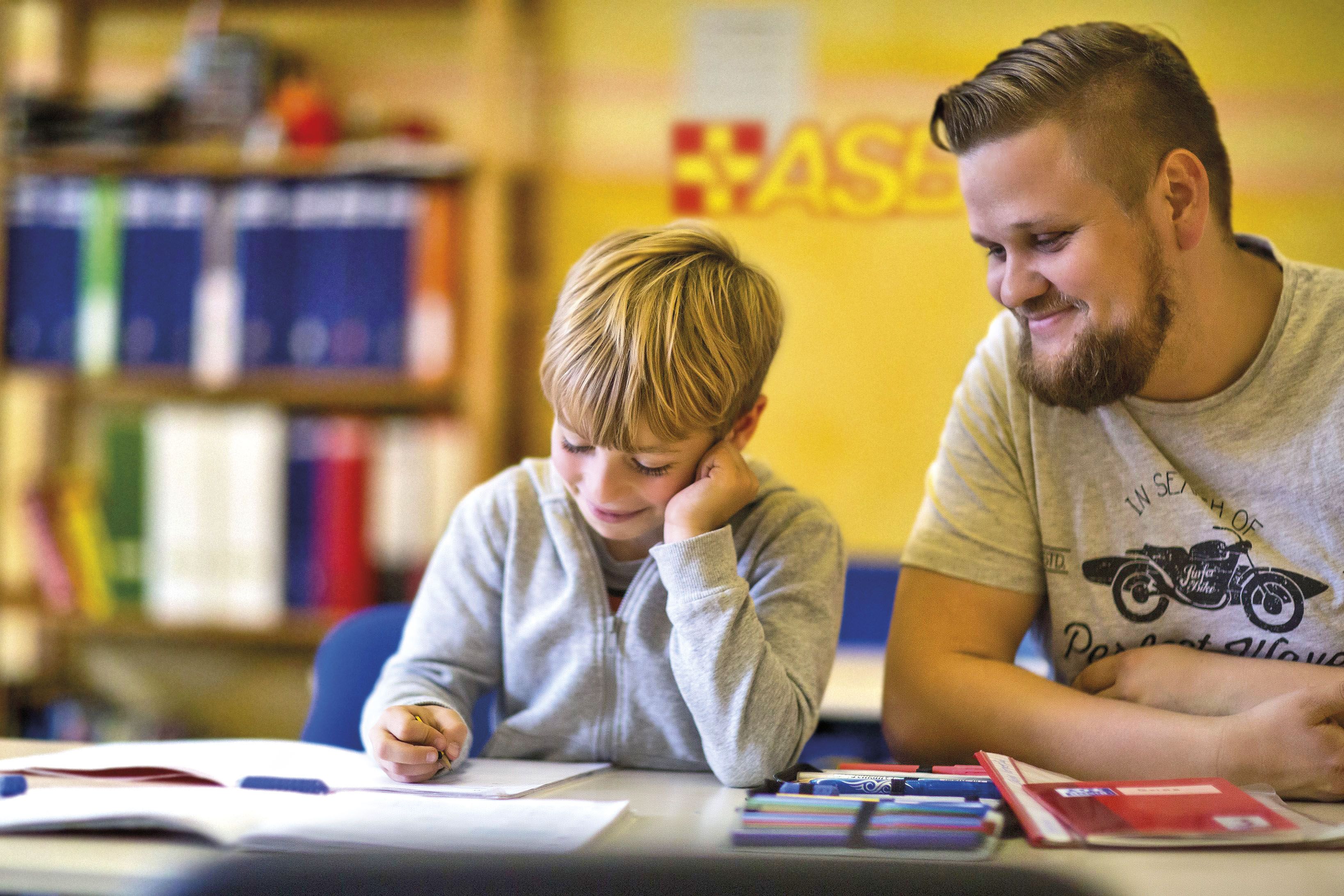Ein junger, freundlich lächelnder Sozialarbeiter unterstützt einen blonden Jungen bei seinen Hausaufgaben.