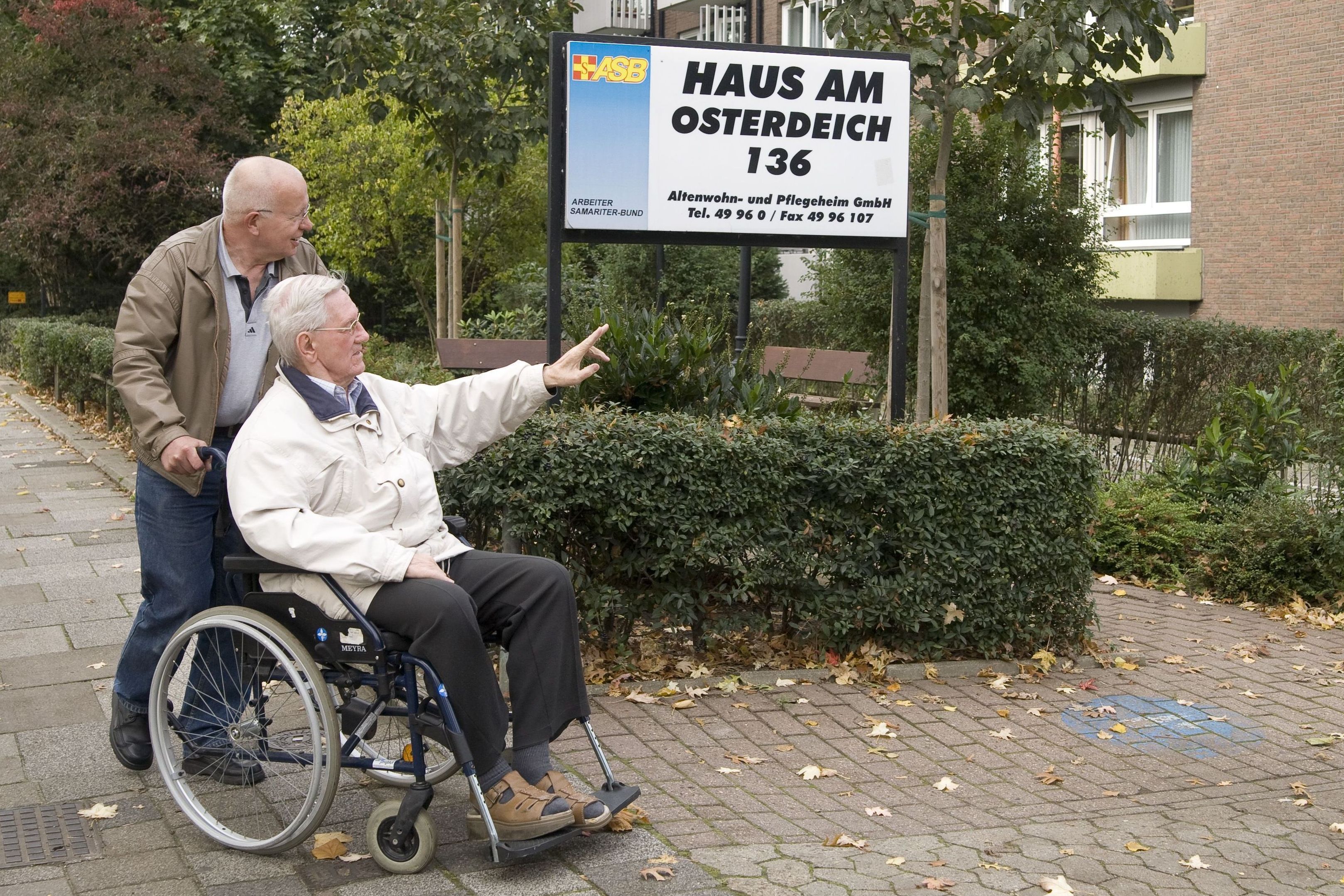 Ein Mann schiebt einen älteren Herrn im Rollstuhl durch einen Park. Gerade kommen sie am Hinweisschild auf die ASB-Seniorenresidenz vorbei