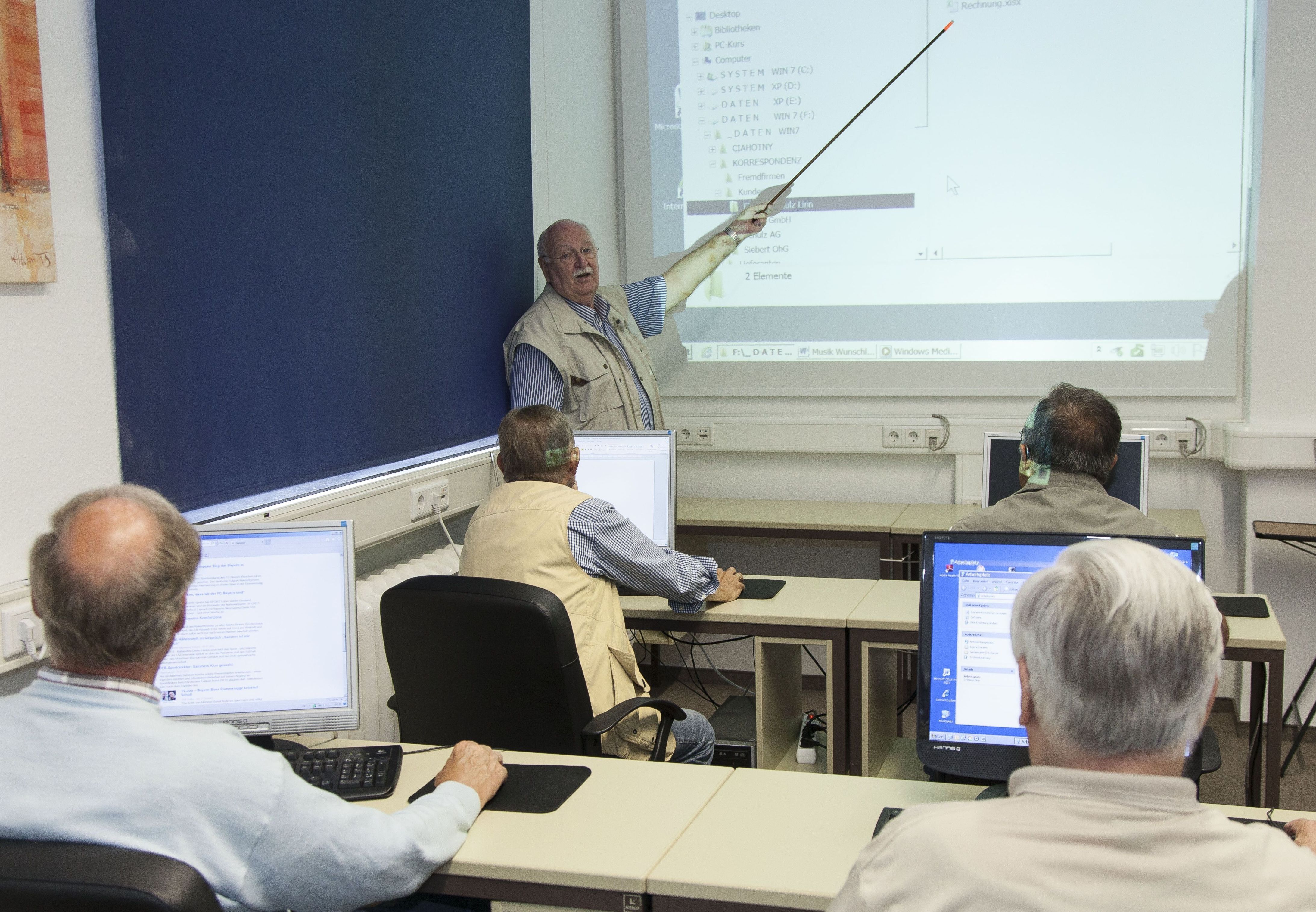 Ein älterer Mann erklärt einer kleinen Gruppe von Senioren mit einem Zeigestock die an die Wand projezierte Verzeichnisstruktur auf einem Computer.