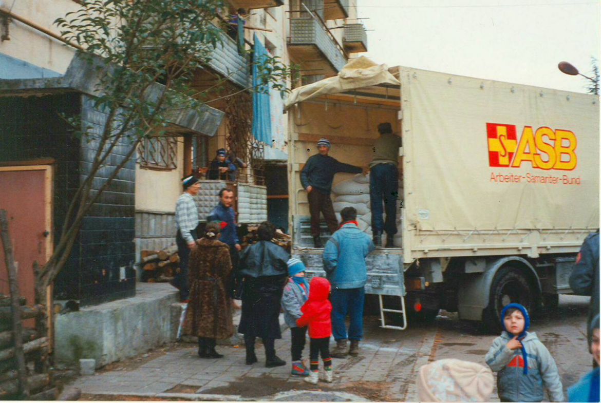 Männer, Frauen und Kinder stehen vor einem ASB-LKW, der Säcke geladen hat. (1995-96)